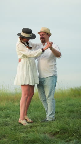 a couple dances together on a grassy hill, with the man wearing a white shirt, hat, and jeans, while the woman is dressed in a white dress and black hat. the serene outdoor setting captures their joy