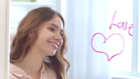 young woman writing by lipstick word love on mirror with heart in bathroom