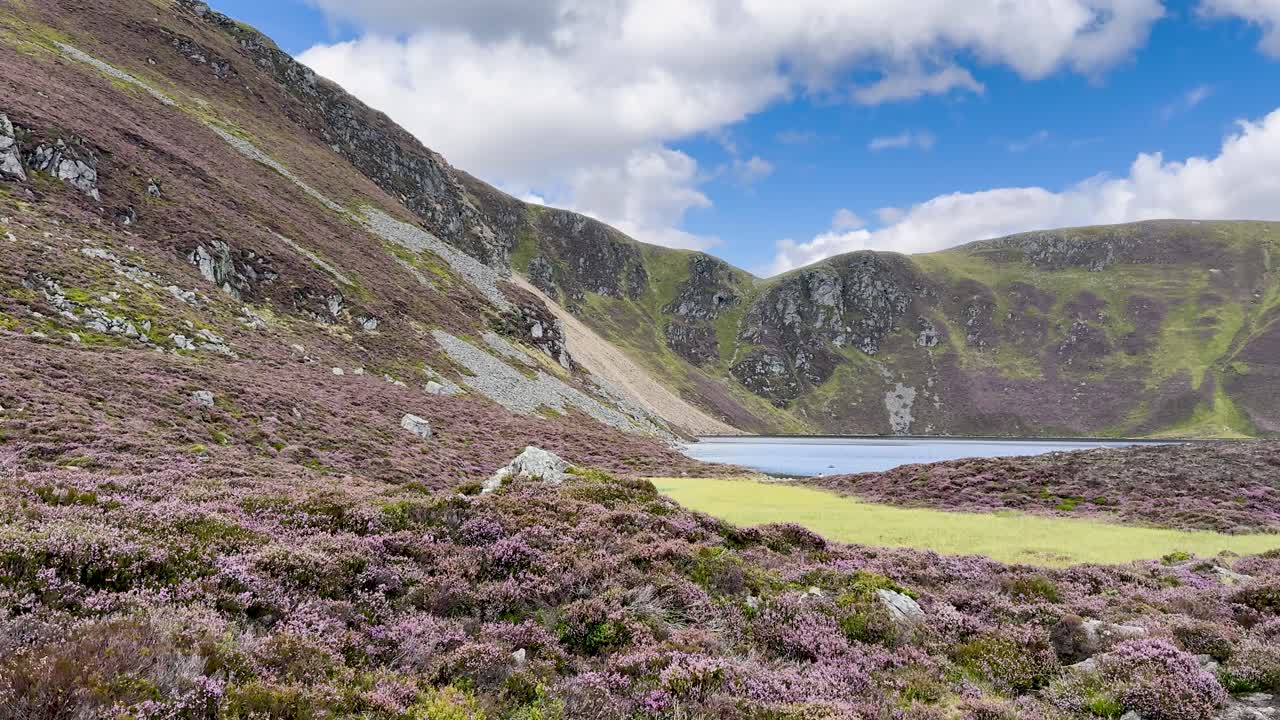 Wide panning shot reveals Loch Brandy surrounded by heather, rocky slopes, and grassy hills under bright daylight with scattered clouds