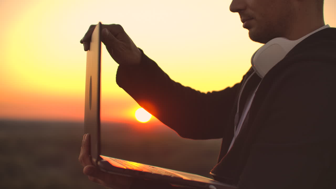un hombre con grandes auriculares blancos escribe con los dedos en el teclado de una computadora portátil de pie en el techo de un edificio al atardecer contra el fondo de la ciudad.
