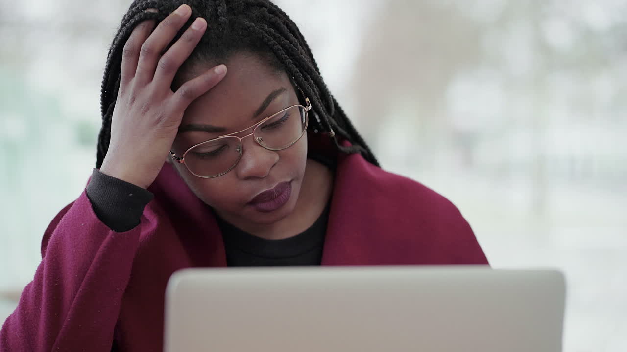 Afro-American attractive young girl with plump rose lips and braids in aviator eyeglasses wearing rose coat working on laptop outside