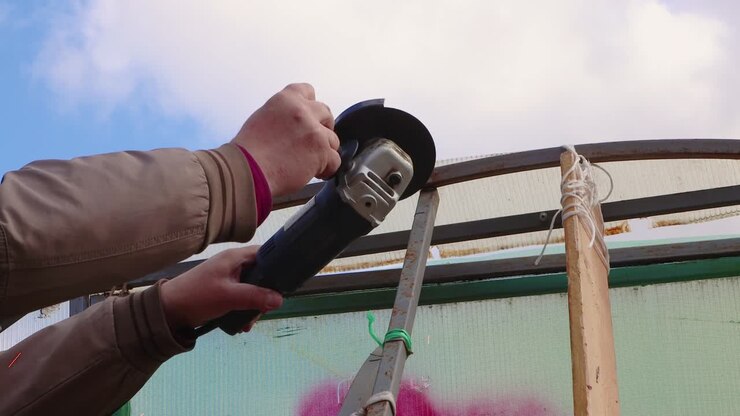 A man with an angle grinder cuts a rusty metal greenhouse against a blue sky.