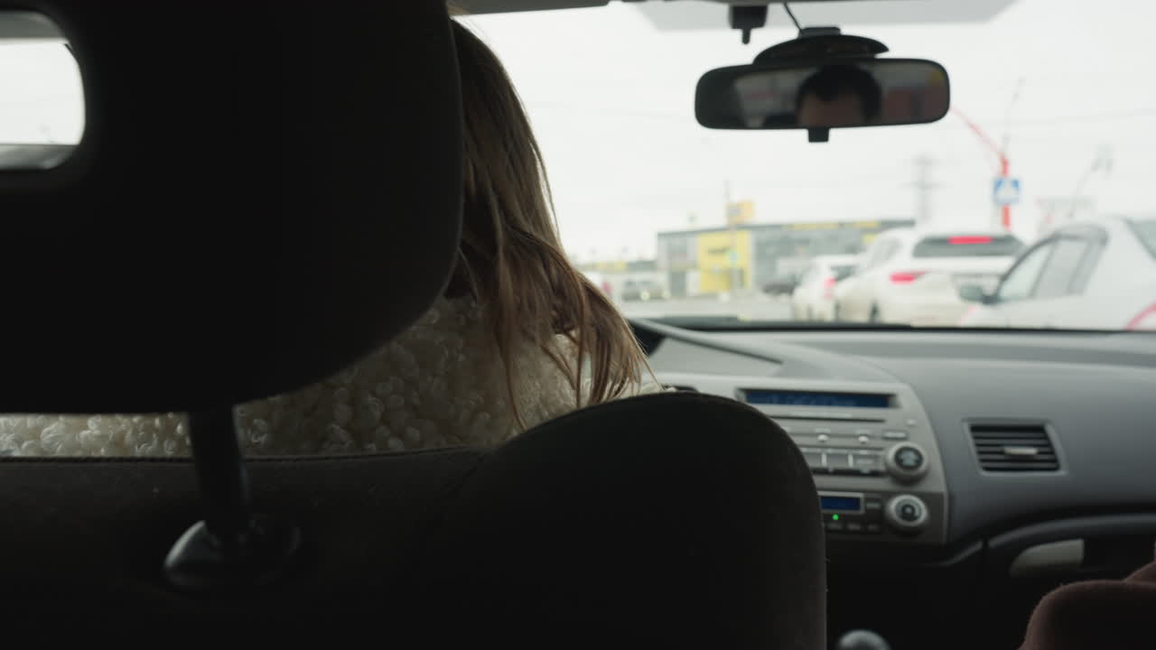 Rear view of woman wearing fluffy sweater driving car during snowy day with parked white vehicle visible through windscreen, showing winter landscape, frozen road, and calm traffic conditions ahead