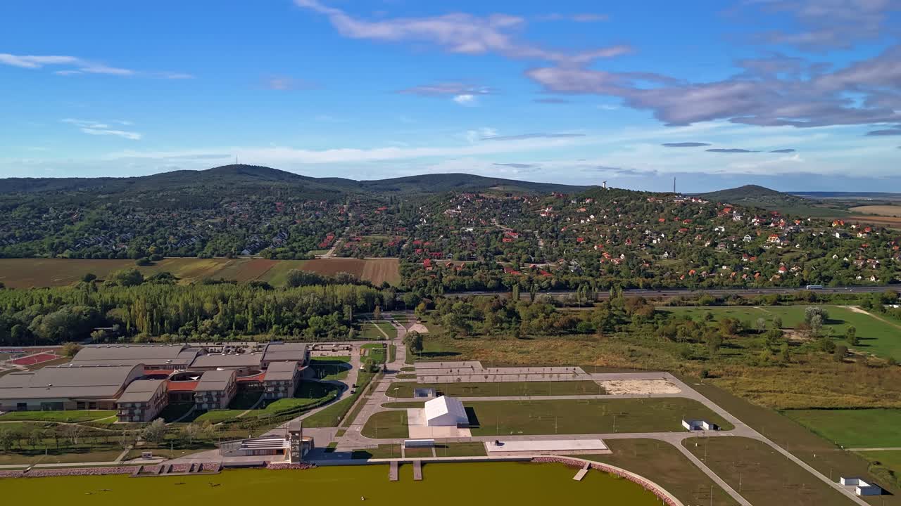 Forward-moving drone hyperlapse approaching the National Kayak-Canoe Academy and M7 highway at Lake Velence, Hungary, with green hills and autumn countryside in the background