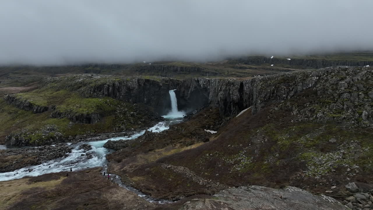 cascada folaldafoss: toma aérea con movimiento de acercamiento a la hermosa cascada y por encima pasa una mujer vestida de negro que admira la belleza de la cascada