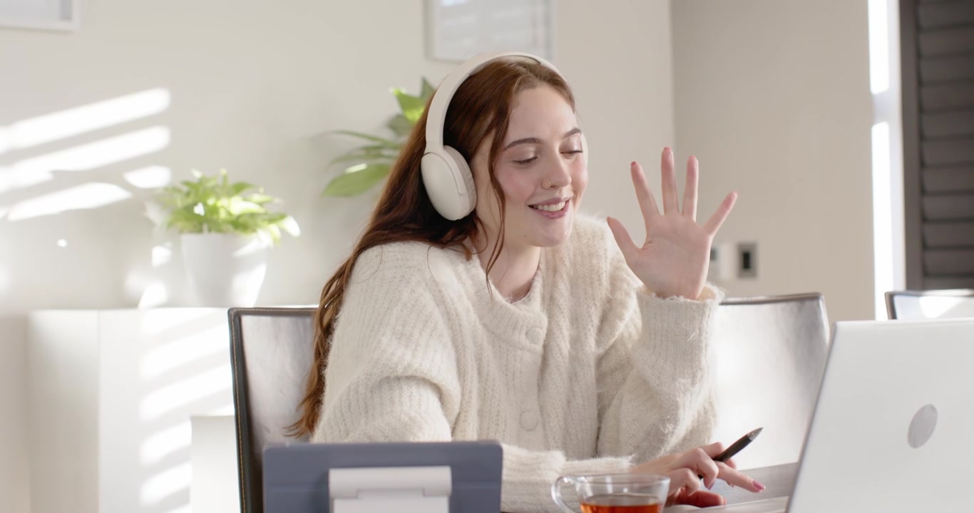 Woman in 20s at home wearing headset, joining online meeting, gesturing with pen near laptop
