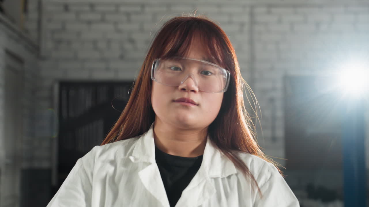 실험실 코트를 입은 중국 여성은 자동차 작업장에서 보호 안경을 조정하는 동안 태블릿을 들고 있습니다. 밝은 빛은 뒤에서 빛나고, 그녀의 집중된 표정에 빛을 습니다.