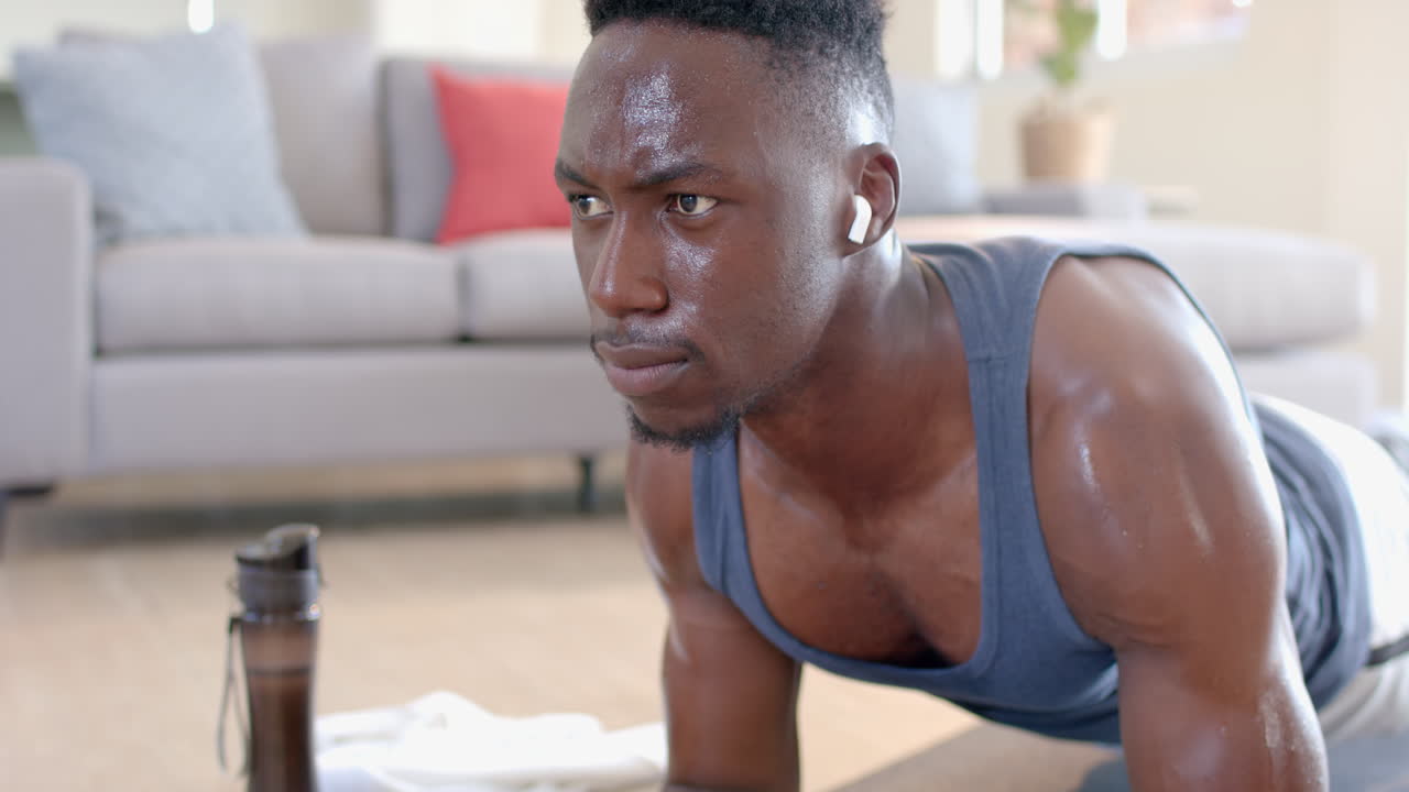 Tired african american man doing plank and using headphones in sunny living room, slow motion