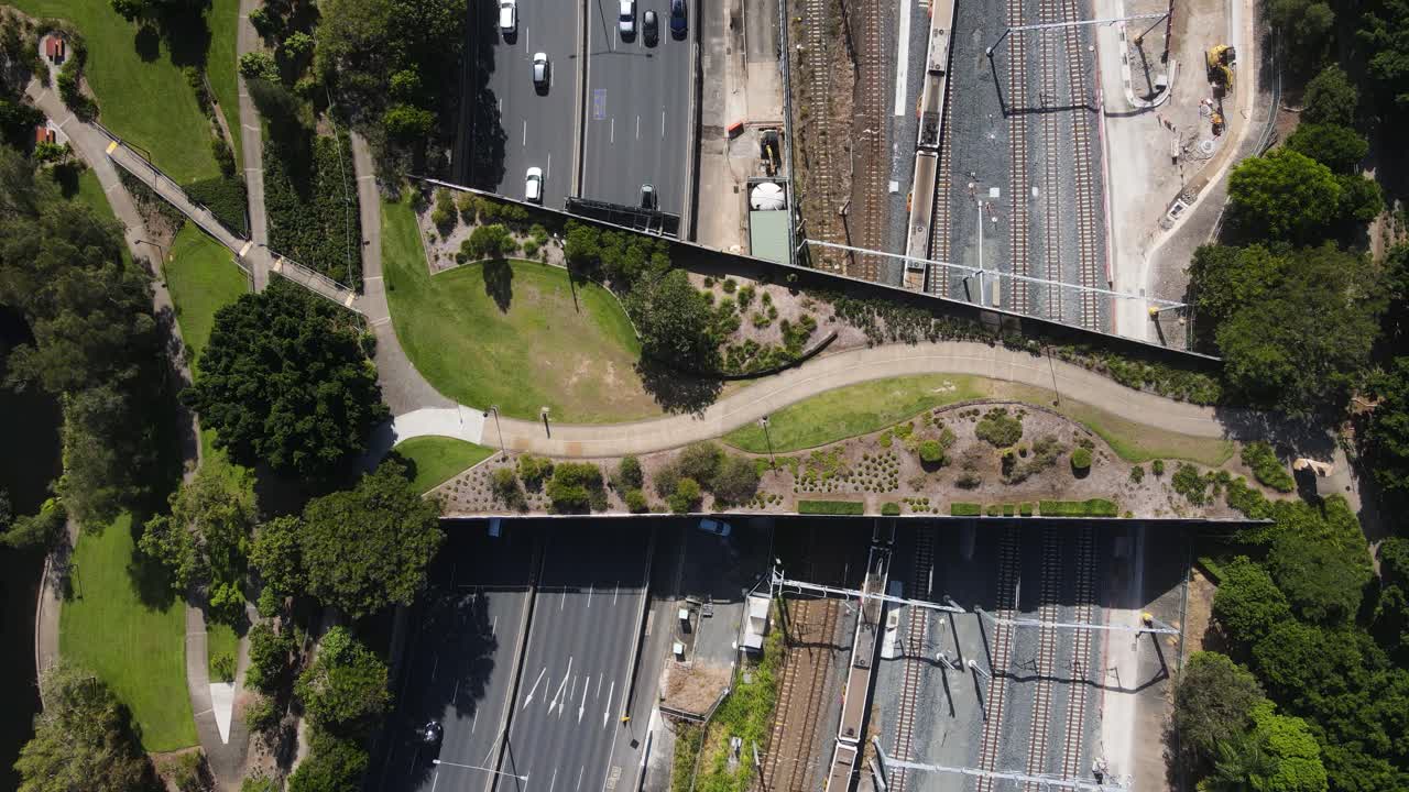 Trains and vehicles pass under an urban city green bridge connecting parkland to a metropolitan city landscape. Aerial view