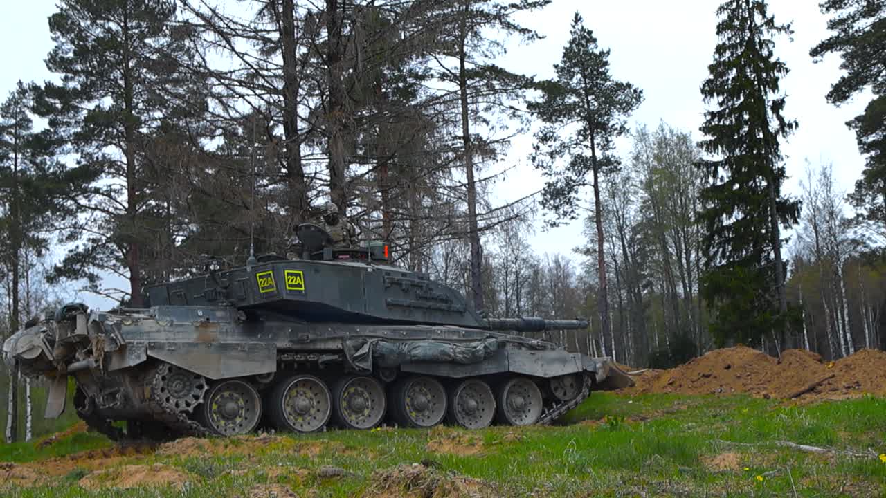Rare footage of a British army or military Challenger 2 4034 tank with a bulldozer plough reversing away from infantry trenches after a battle during a cloudy day in a pine forest with white sky.