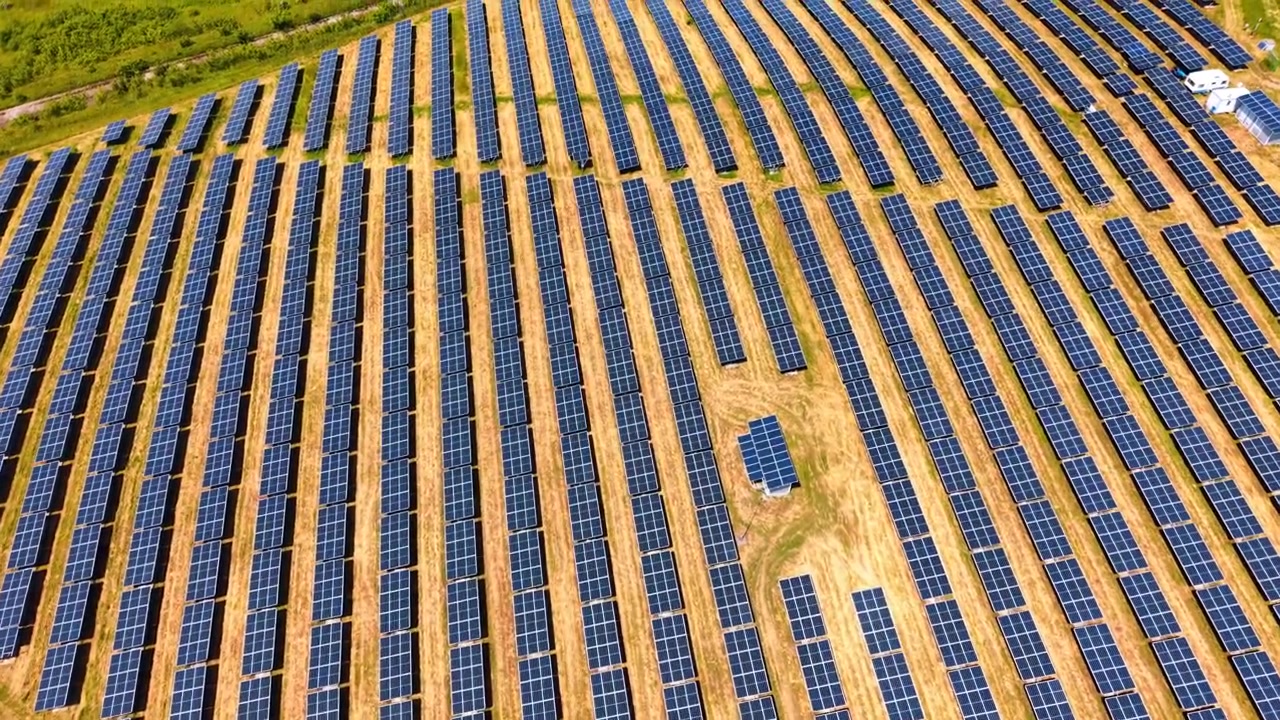File diagonali di pannelli solari su terreno arido. Array fotovoltaici simmetrici su campo d'erba gialla, piccolo edificio bianco, vista aerea, disposizione geometrica.