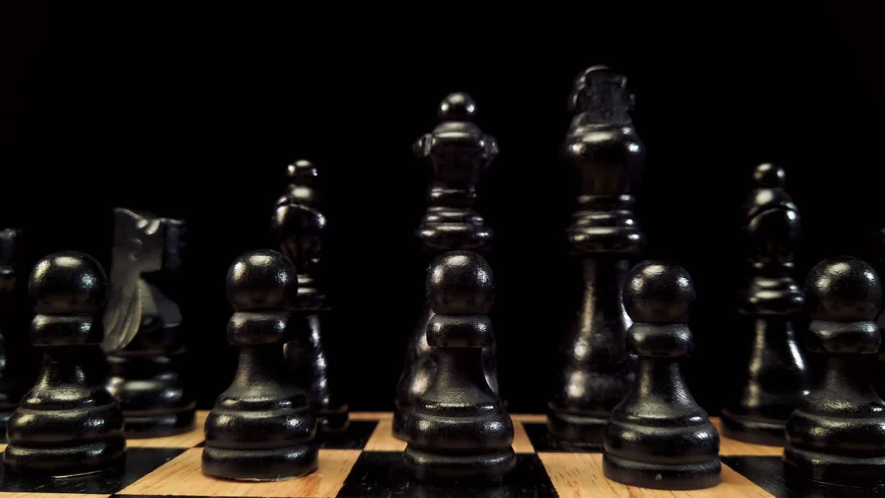 Truck shot from right to left of black chess pieces standing on a chessboard on a black background