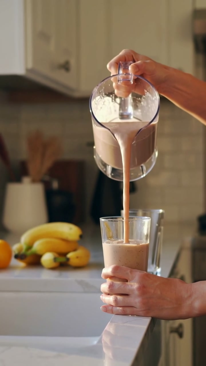 Vertical video: Tilting pitcher, hands pouring tan shake into glass on kitchen counter to drink