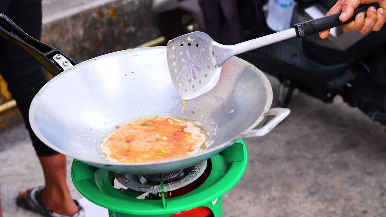 A street vendor prepares an omelet in a wok on a portable stove, showcasing traditional cooking techniques