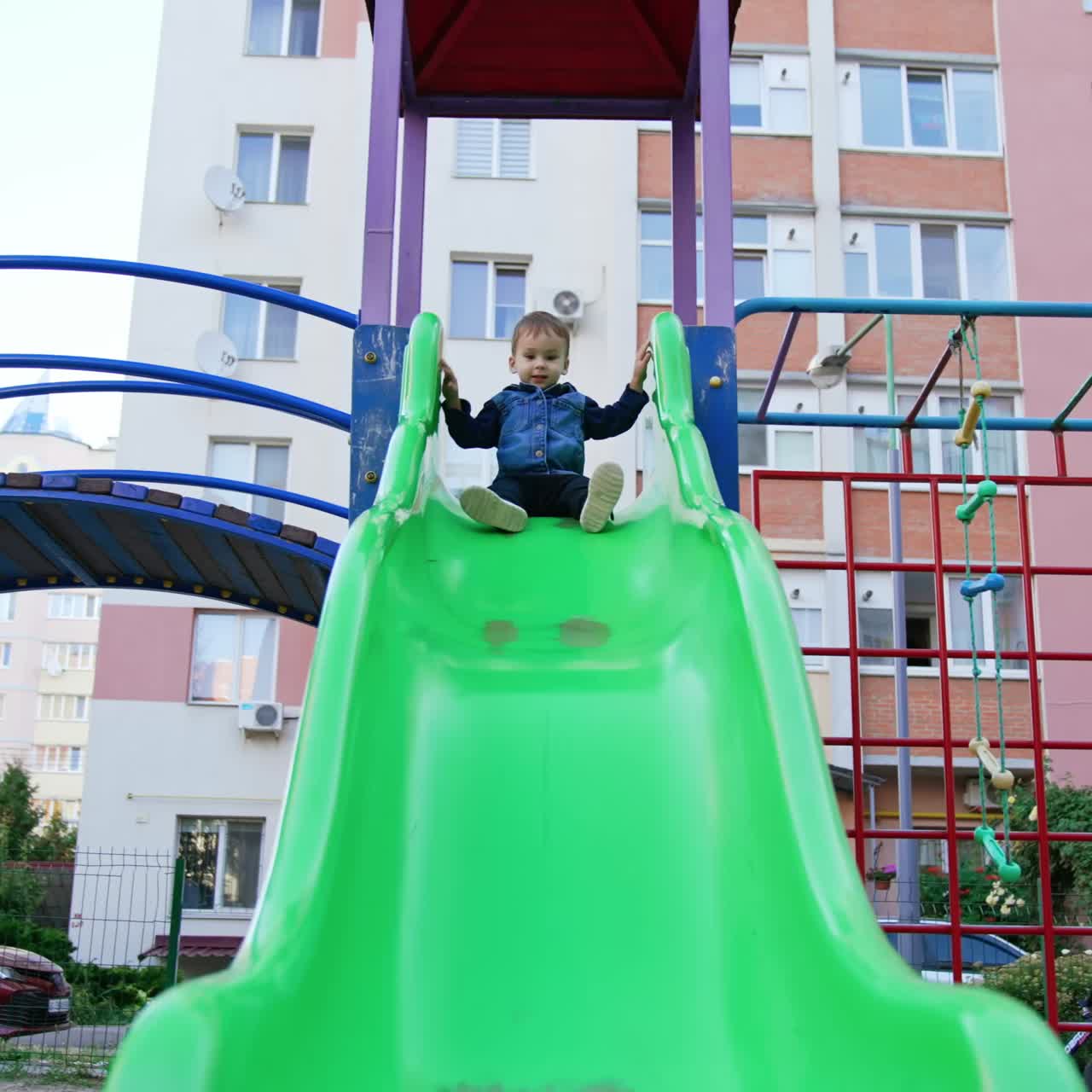 Little kid sits on the big green slide. Happy kid slides down approaching camera. Low angle view