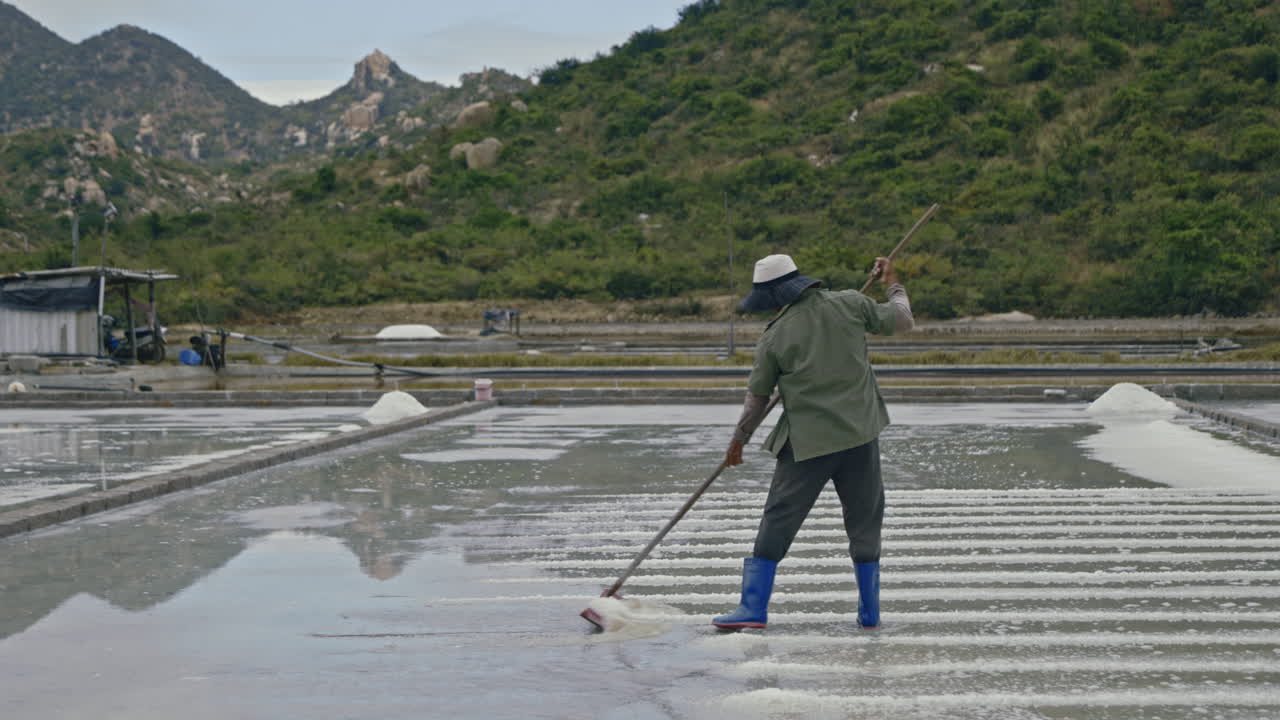 A person raking salt in a traditional salt field with mountains in the background