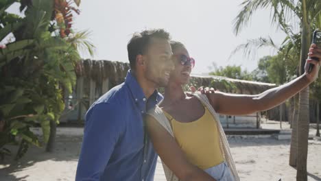 happy diverse couple smiling and taking selfie with smartphone on sunny beach, in slow motion