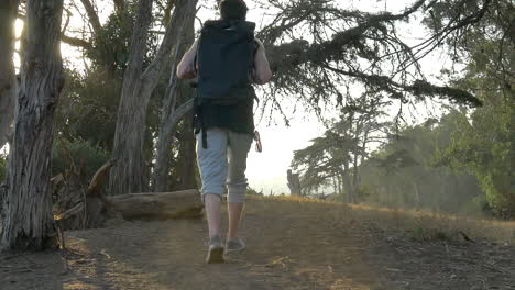 young man traveler wearing a large black backpack walking in slow motion along the ocean bluffs during a golden sunset in santa barbara, california