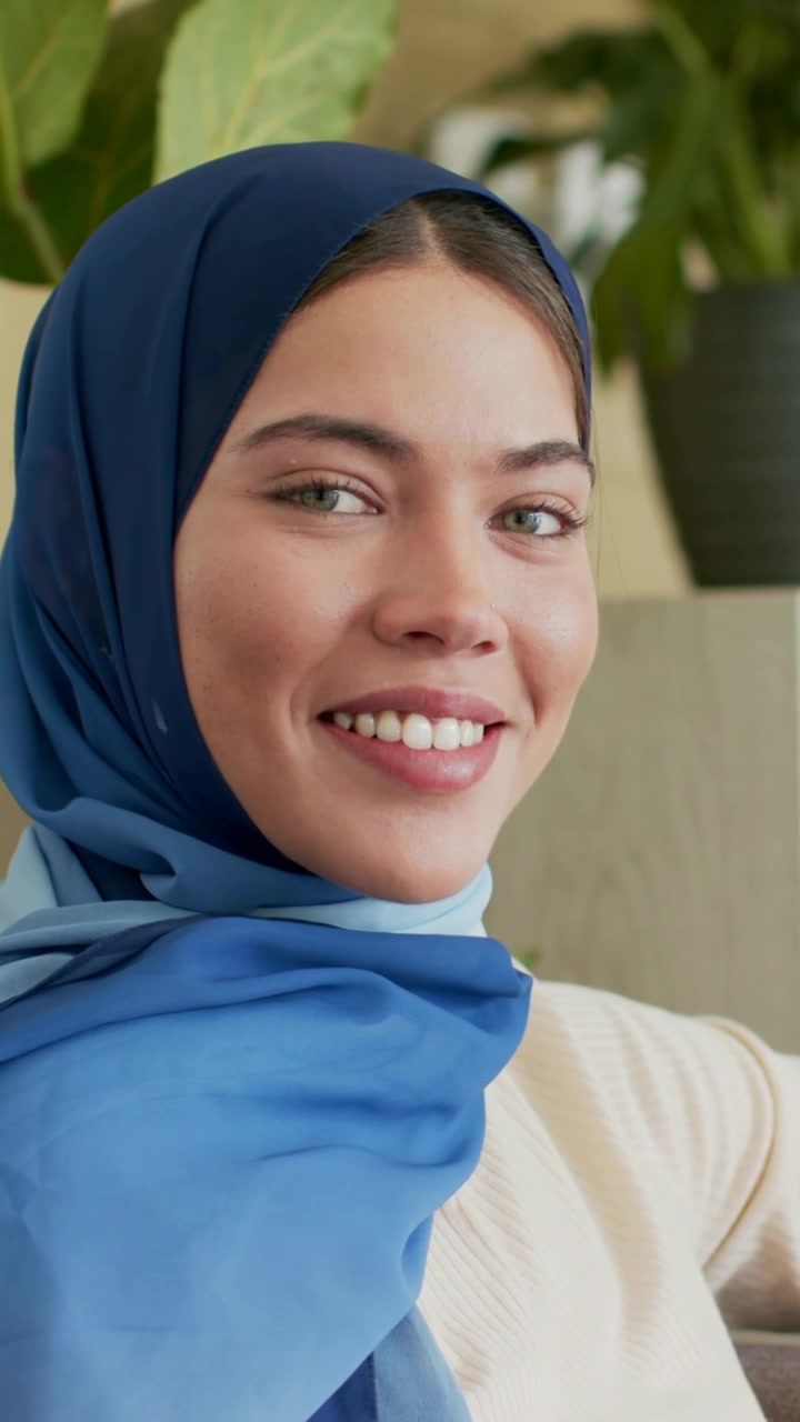 Vertical video: Recording, woman wearing blue hijab smiling, turning head on sofa with plant