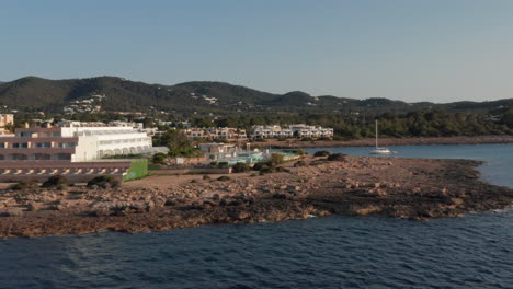 Vista-Aérea-Del-Agua-Del-Océano-Silenciosa-Y-Tranquila-Que-Fluye-Tocando-La-Orilla-Con-Resorts-Y-Villas-En-Una-Casa-De-Vacaciones-Con-Yates-Y-Barcos-Estacionados-En-Ibiza-En-España
