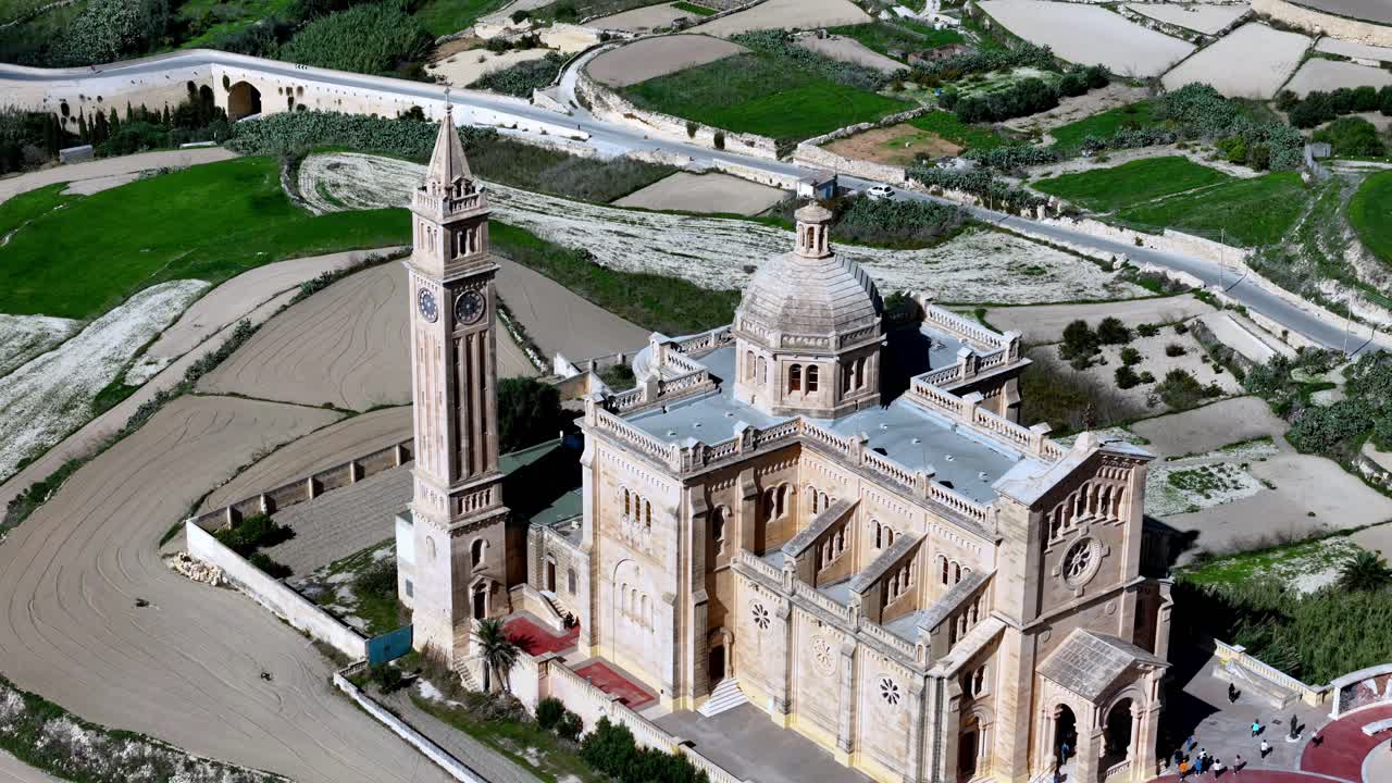 Gozo Malta'daki Ta Pinu Bazilikası'nın, parlak bir bahar gününde, açık mavi güneşli bir gökyüzünün altında yemyeşil tarlalar ve teraslı araziler önünde ikonik kireçtaşı mimarisini sergileyen havadan görünümü.