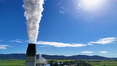 a smoke stack releases steam into a bright blue sky over a rural landscape, highlighting industrial impact on the environment