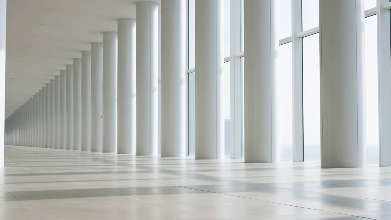 Orderly rows of white pillars recede toward a distant vanishing point beside large window panels, while diffuse daylight creates soft shadows and reflections on clean flooring.