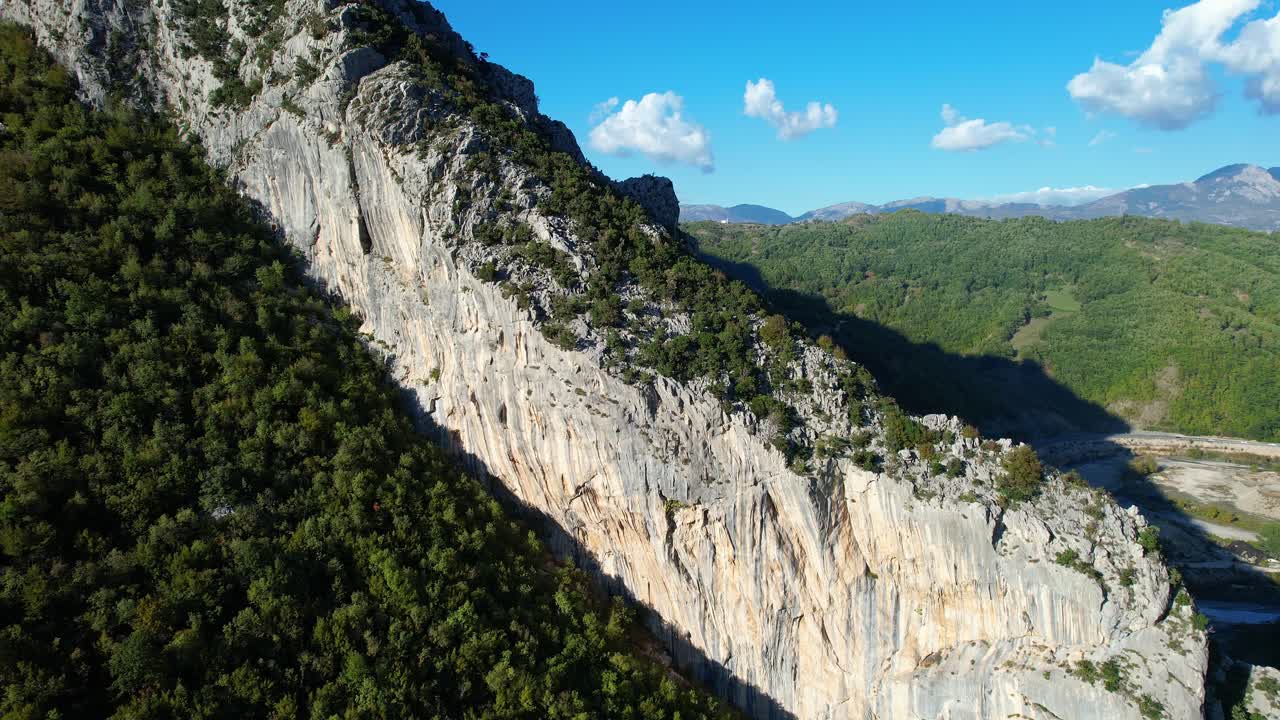 Panoramic shot of a climber descending the Brar vertical rock wall, near Tirana, with a green mountainous valley background and blue sky