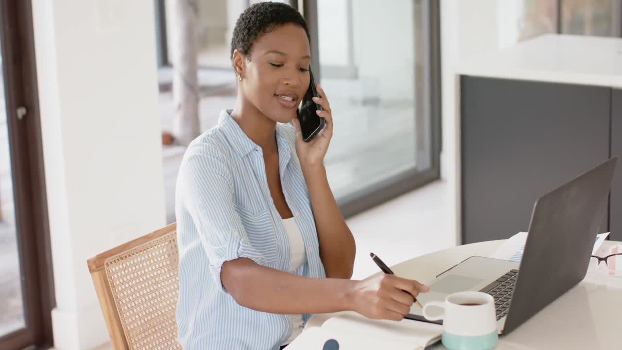 African american woman sitting at table talking on smartphone and taking notes, slow motion