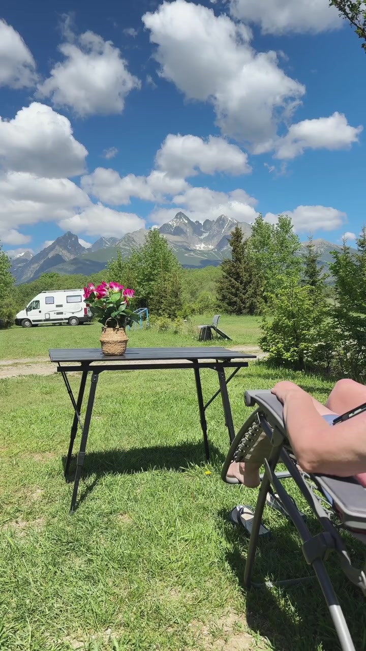 Nainen rentoutuu aurinkotuolissa slovakialaisella leirintäalueella, josta avautuu panoraamanäkymä Tatravuorille, nauttien rauhallisesta ulkoilulomasta.