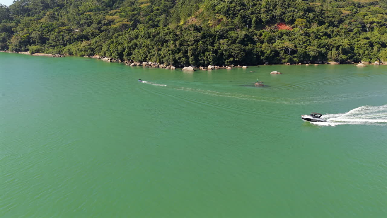 Aerial drone shot of a sleek, luxury yacht speeding across the emerald green waters of Balneário Camboriú, Brazil. A symbol of wealth and exclusive summer vacations