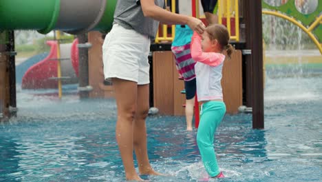 mother and young child girl have fun together in aqua park playground, kid jumping high splashing shallow water holding mom's hands - slow motion