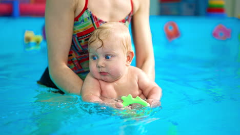 adorable caucasian baby with blond hair is learning to swim. coach moves the kid in the water while child is focused on toy.