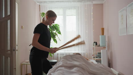 massage therapist in black outfit smiling while using bamboo massage broom on client lying face down under white blanket in softly lit spa room with indoor plant and sheer curtains