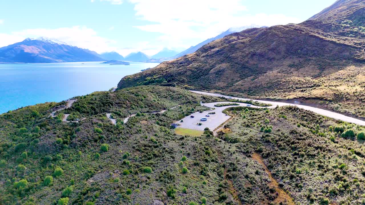 Drone glides above winding mountain road, parking lot, and lake under bright daylight, wide perspective