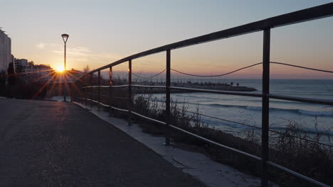 Der-Zeitraffer-Erfasst-Den-Sonnenaufgang-Auf-Der-Straße-In-Der-Nähe-Des-Geländers,-Das-Die-Küste-Vom-Meer-Trennt