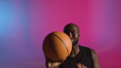 Retrato-De-Estudio-De-Un-Jugador-De-Baloncesto-Masculino-Girando-La-Pelota-En-El-Dedo-Contra-Un-Fondo-Iluminado-De-Color-Rosa-Y-Azul