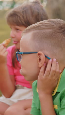 el niño de camisa verde se mete todo su bocadillo en la boca mientras su hermana de camisa rosa disfruta de la suya más suavemente, con la madre observando al aire libre en el campo abierto durante el picnic familiar