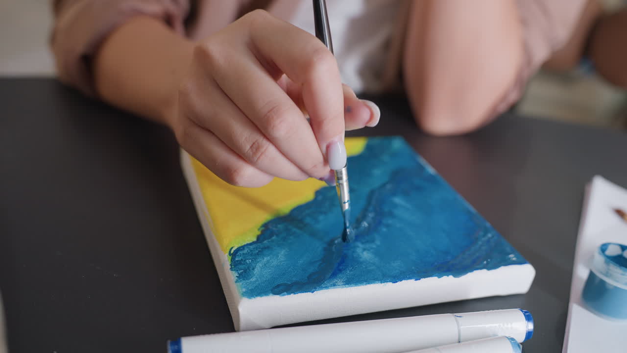 Primer plano de un pincel cargado con esmalte azul aplicando un trazo controlado sobre un lienzo texturizado, con una base amarilla asomando por debajo, rotuladores extendidos, movimiento concentrado de la mano creando un efecto de capas
