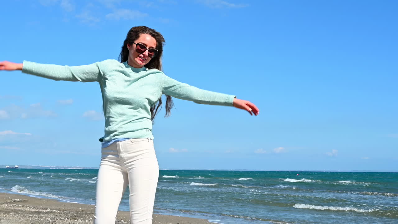 Cheerful woman in sunglasses and casual clothes jumping with joy on the sandy beach of Larnaca, Cyprus, with blue sea waves in the background
