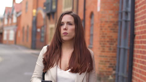 Woman-Outdoors-Walking-To-Work-Along-City-Street-Lined-With-Houses-Looking-At-Mobile-Phone