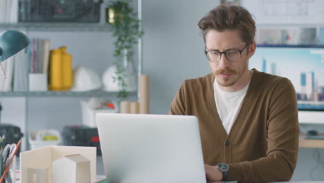 male architect in office working at desk on laptop taking call on mobile phone