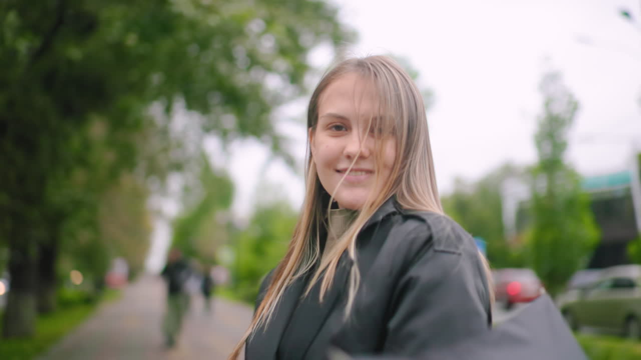 Beautiful lady spinning an umbrella outdoors wearing black coat on city street looking sideways with smile during rainy autumn day with green trees and urban background creating natural candid lifestyle