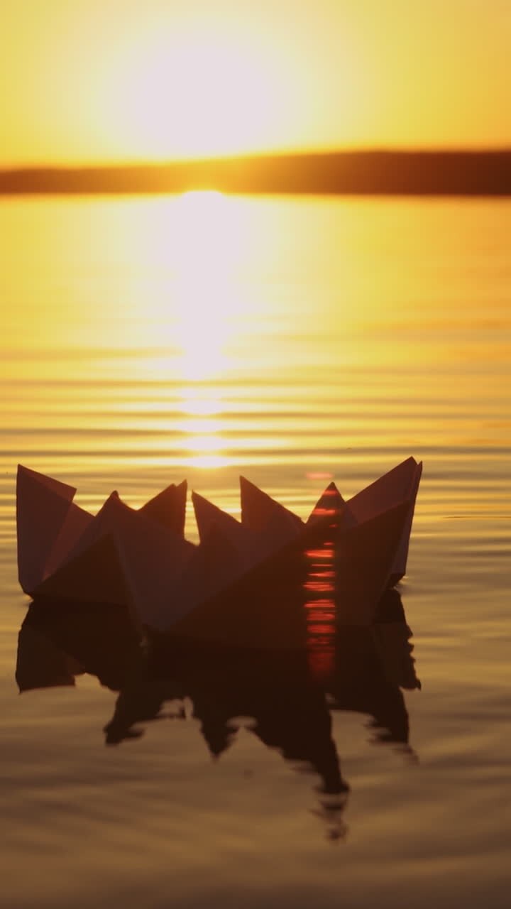 Female's hand putting one paper boat on the water and it joins to other ones at sunset. New origami ship adding to the rest during beautiful sunset with the sun reflection in the lake Vertical video