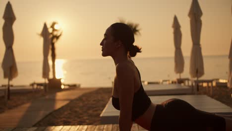 shot close up of a brunette girl with tied hair in a black sports summer uniform doing the cobra exercise and doing yoga on a sunny beach in the summer. golden hour, golden sunrise