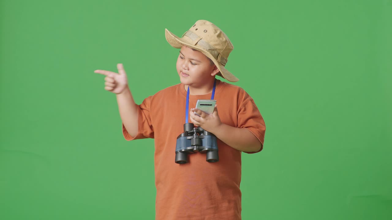 모자와 안경을 쓴 아시아 관광 소년은 스마트폰을 보고 미소 짓고 녹색 화면 배경에서 을 가리키고 있습니다. 소년 연구원은 여행, 관광, 모험 개념의 무언가를 조사합니다.