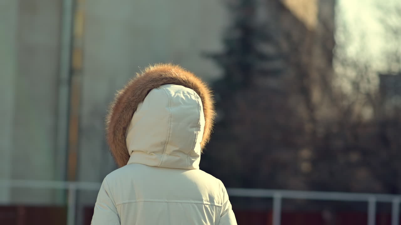 A person wearing a white coat with a fur hood enjoys a peaceful winter stroll in the city. Warm sunlight brightens the surroundings, creating a calm atmosphere