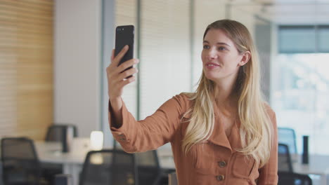 mujer de negocios teniendo una conversación de video en una llamada de teléfono móvil de pie en una oficina moderna de plan abierto