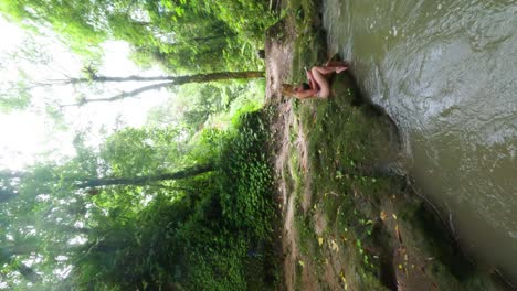 Vertical-FPV-Drone-panning-shot-of-beautiful-blond-woman-relaxing-in-the-nature-of-Bali-with-a-large-waterfall-in-the-background