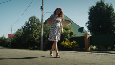 remote worker walking confidently on sunny street as she removes sunglasses and places them on head, wearing floral dress with breeze lifting hemline, framed by trees, buildings, and clear sky